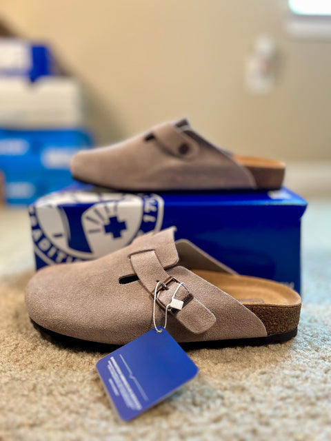 Beige clogs with a blue tag on a carpeted floor, with a blue shoebox in the background.