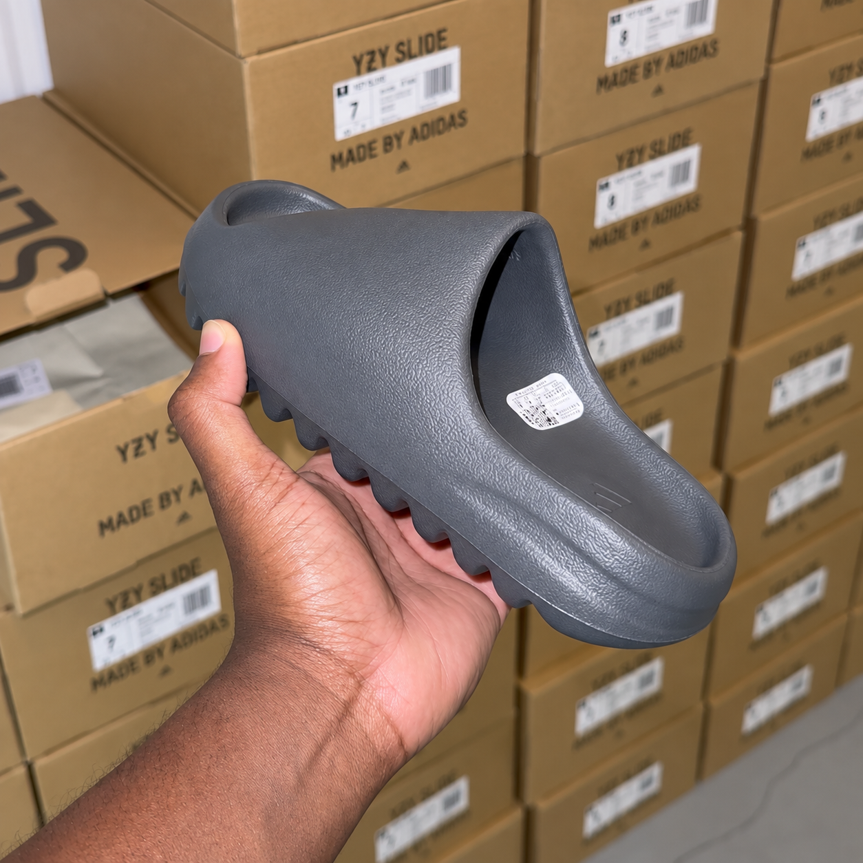 Charcoal Yeezy Slide held in hand in front of stacked shoe boxes